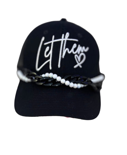 Black cap with 'Let them' text on a stand against a white background