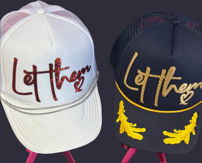 Two hats with 'Let them' text on stands against a blurred background