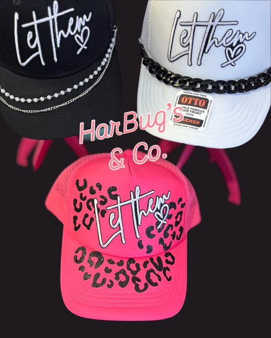 Three hats with 'Let them' text, one pink with leopard print, on a dark background.